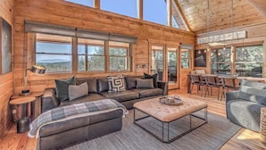 A cozy cabin interior with a large sofa, a coffee table, and a dining area. - Tranquility Ranch - Hot Tub | Pool Table | Fire Pit in Private Yard (Mineral Bluff)