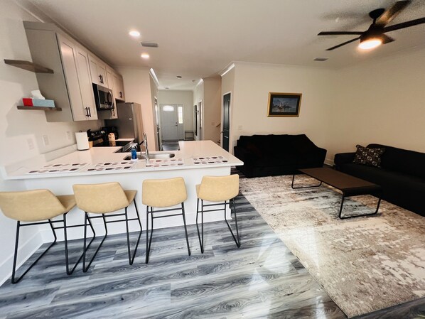 Una cocina moderna con una isla central, un comedor con cuatro sillas, un sofá negro y un ventilador de techo.