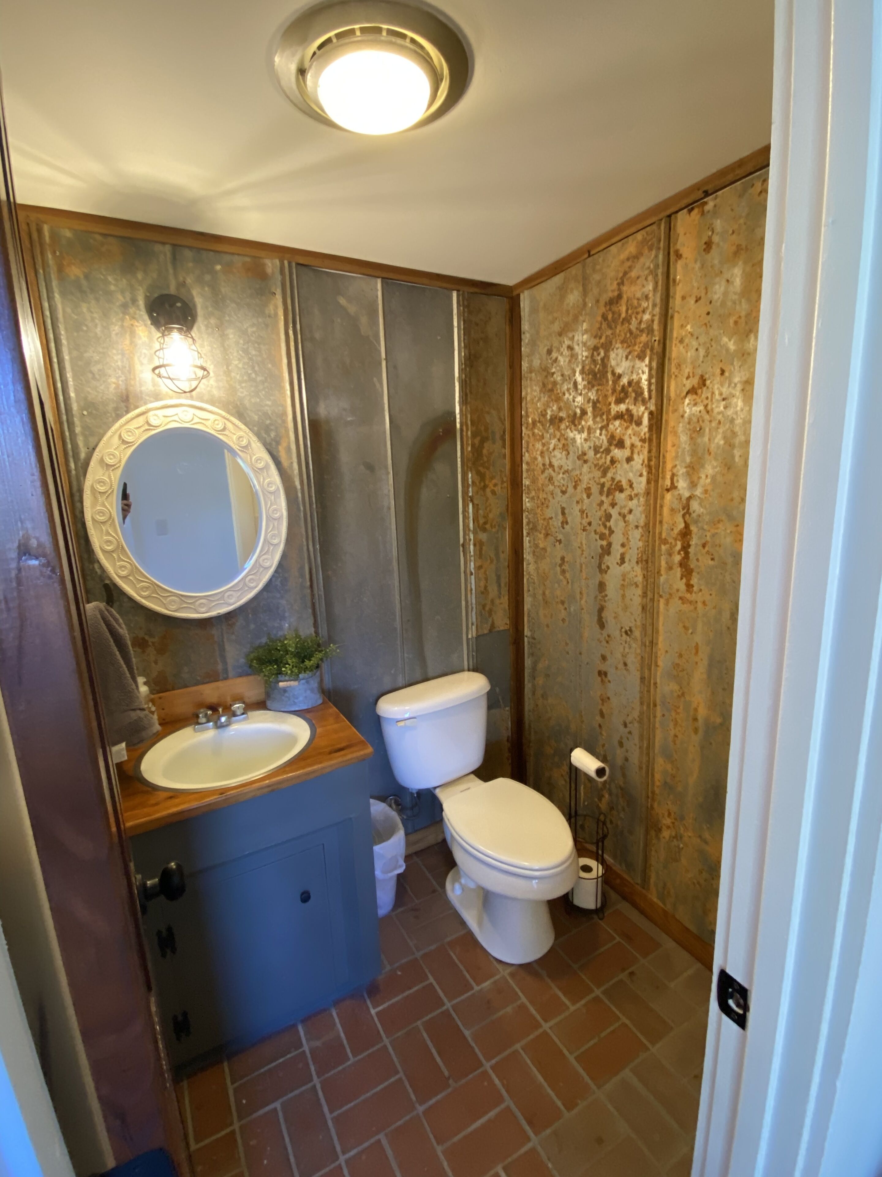 A bathroom with a wooden vanity, a round mirror, a toilet, and a trash bin.