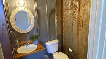A bathroom with a wooden vanity, a round mirror, a toilet, and a trash bin.