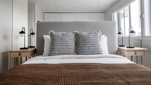A modern bedroom with a large bed, two bedside tables with lamps, a grey headboard, and a window with a view. - Modern Chelsea 1BR w/ Balcony nr shops, by Blueground (London)
