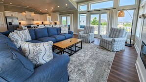 A spacious living area with a large blue sectional sofa, a wooden coffee table, and a gray rug. There are two upholstered chairs and a floor lamp. The room has large windows with curtains, a ceiling fan, and a kitchen in the background.