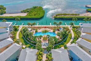 Luchtbeeld van een resort met een centraal zwembad, omgeven door weelderige begroeiing en palmbomen, met een strand en een waterpartij op de achtergrond.