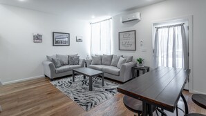 Ein modernes Wohnzimmer mit einer grauen Sofagarnitur, einem Holztisch und einem Esstisch. An der Wand befinden sich gerahmte Bilder, ein Fenster mit Vorhängen und eine Klimaanlage.