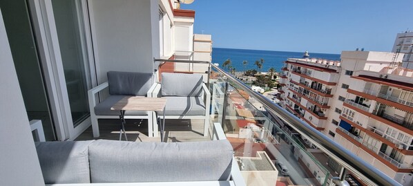 Un balcón con sofá y mesa, con vista a una zona costera con edificios y el mar.