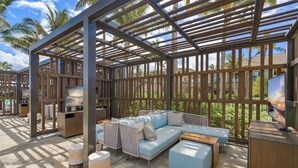 An outdoor lounge area with a wooden pergola, a light blue sofa set, and a television screen.