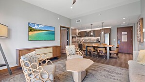 A modern living room with a large flat-screen TV, wicker furniture, and a dining area with a kitchen in the background.