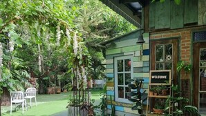 A small, rustic building with a 'WELCOME' sign, surrounded by lush greenery and potted plants. - Ruennakornin (Bang Kruai)