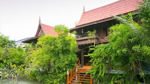 A traditional wooden house with a red roof, surrounded by lush greenery and a small water body. - Ruennakornin (Bang Kruai)