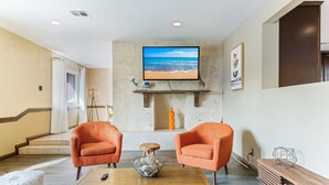 A modern living room with two orange armchairs, a wooden coffee table, a flat-screen TV displaying a beach scene, and a staircase leading to an upper level. - Amazing 6 BRD, Pool, Theater, Billiard, Poker, and Games near the Strip (Las Vegas)