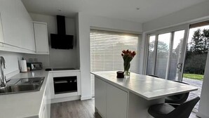 Une cuisine moderne avec des meubles blancs, un îlot central doté de tabourets, un four encastré et une fenêtre avec des stores.