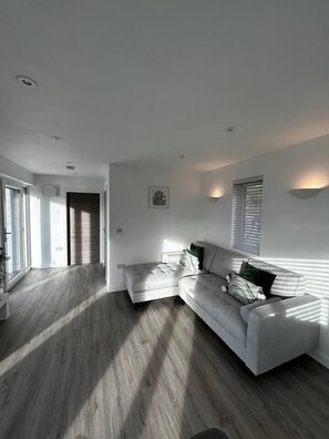 A modern living room with a grey sectional sofa, a flat-screen TV, and a large window providing natural light. - Lowern, a luxury retreat near Menai Bridge (Menai Bridge)