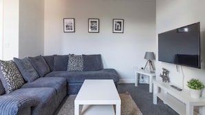 Una sala de estar moderna con un sofá seccional gris, una mesa de centro blanca, un televisor de pantalla plana montado en la pared y una mesita de noche con una lámpara y elementos decorativos.