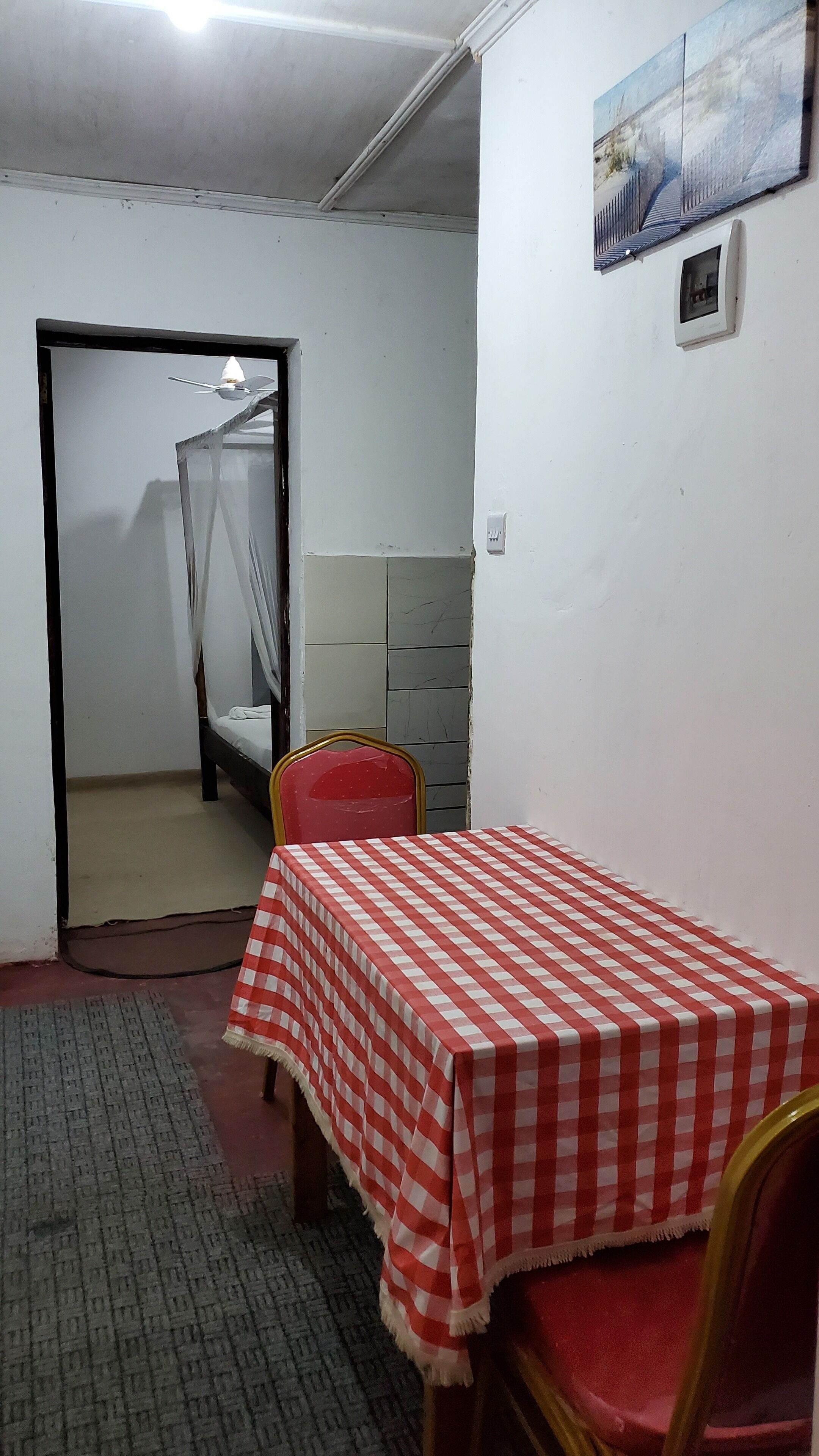 Une chambre à coucher avec un lit, une table recouverte d’une nappe à carreaux, une chaise rouge, une table de chevet, un tableau accroché au mur et une porte donnant sur une autre pièce.