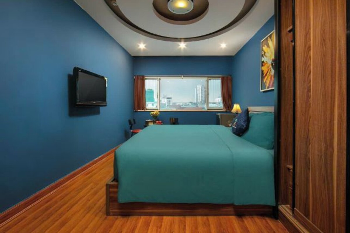 Une chambre avec une couvre-lit turquoise, un téléviseur fixé au mur, une fenêtre avec des rideaux et une vue sur la ville.