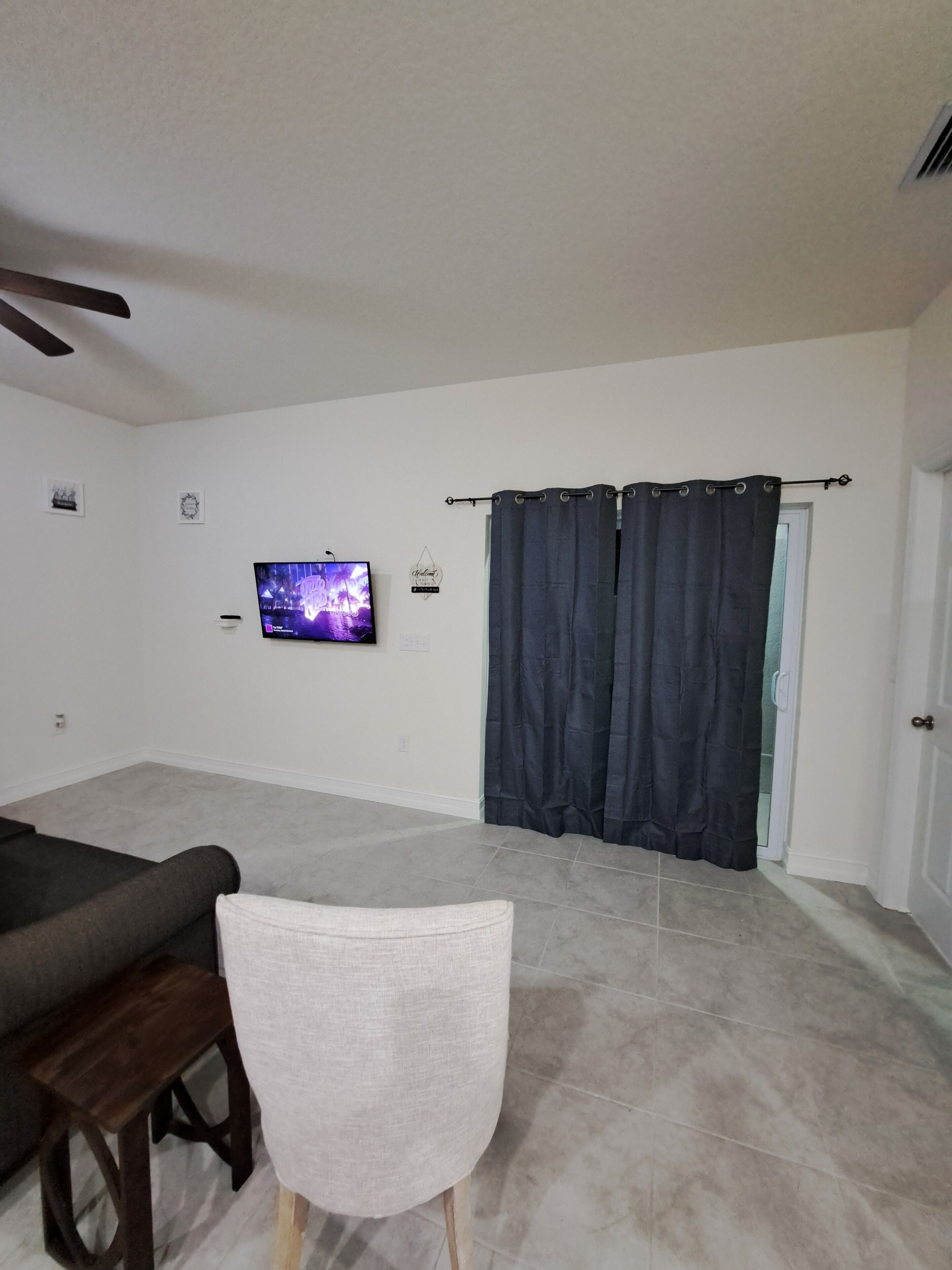 Una sala de estar con un televisor de pantalla plana, un sofá oscuro, una silla blanca y una cortina oscura.