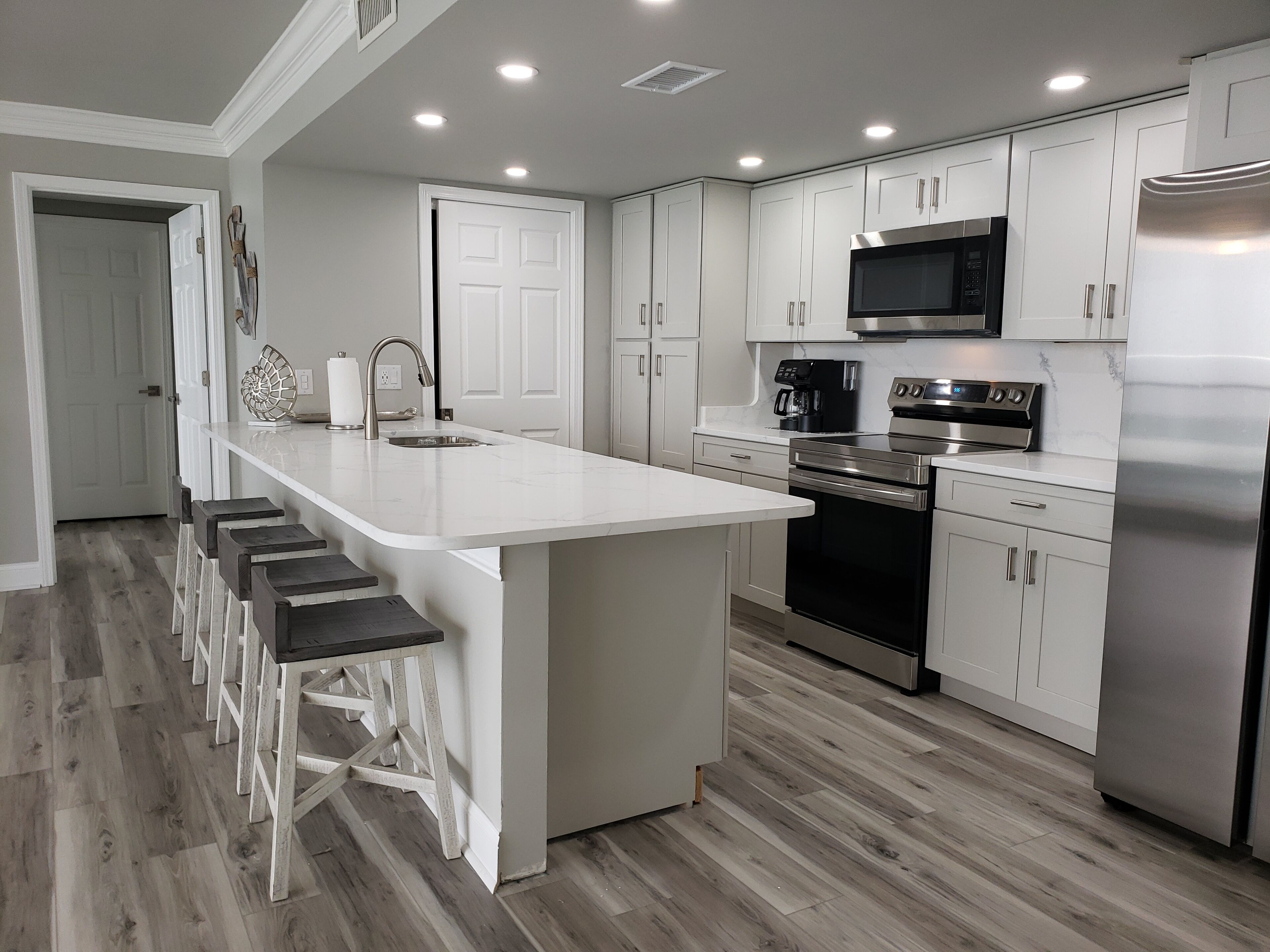 A modern kitchen with white cabinets, a stainless steel refrigerator, a built-in oven, and a microwave. There are bar stools along the kitchen island.