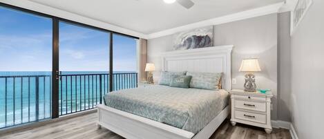A bedroom with a large window overlooking the ocean, a white bed, two bedside tables with lamps, and a ceiling fan.