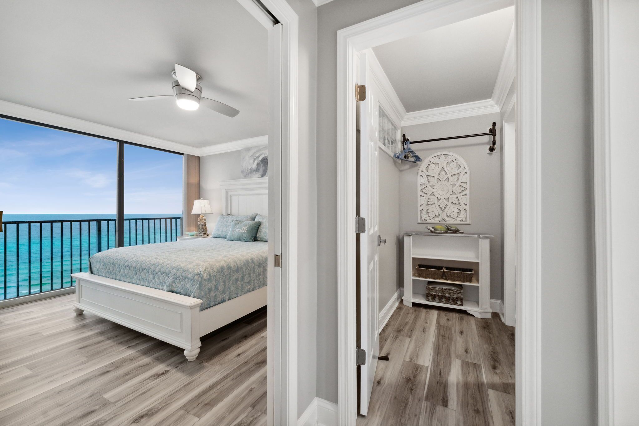 A bedroom with a large bed, a ceiling fan, a view of the ocean, and a small bathroom with a vanity and shelves.