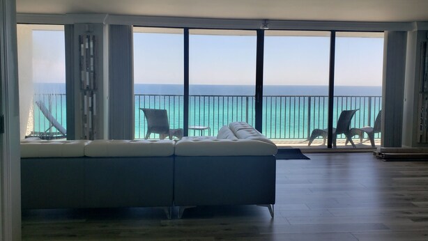 A spacious living room with a large sofa and two chairs, a balcony with a railing, and a view of the ocean.