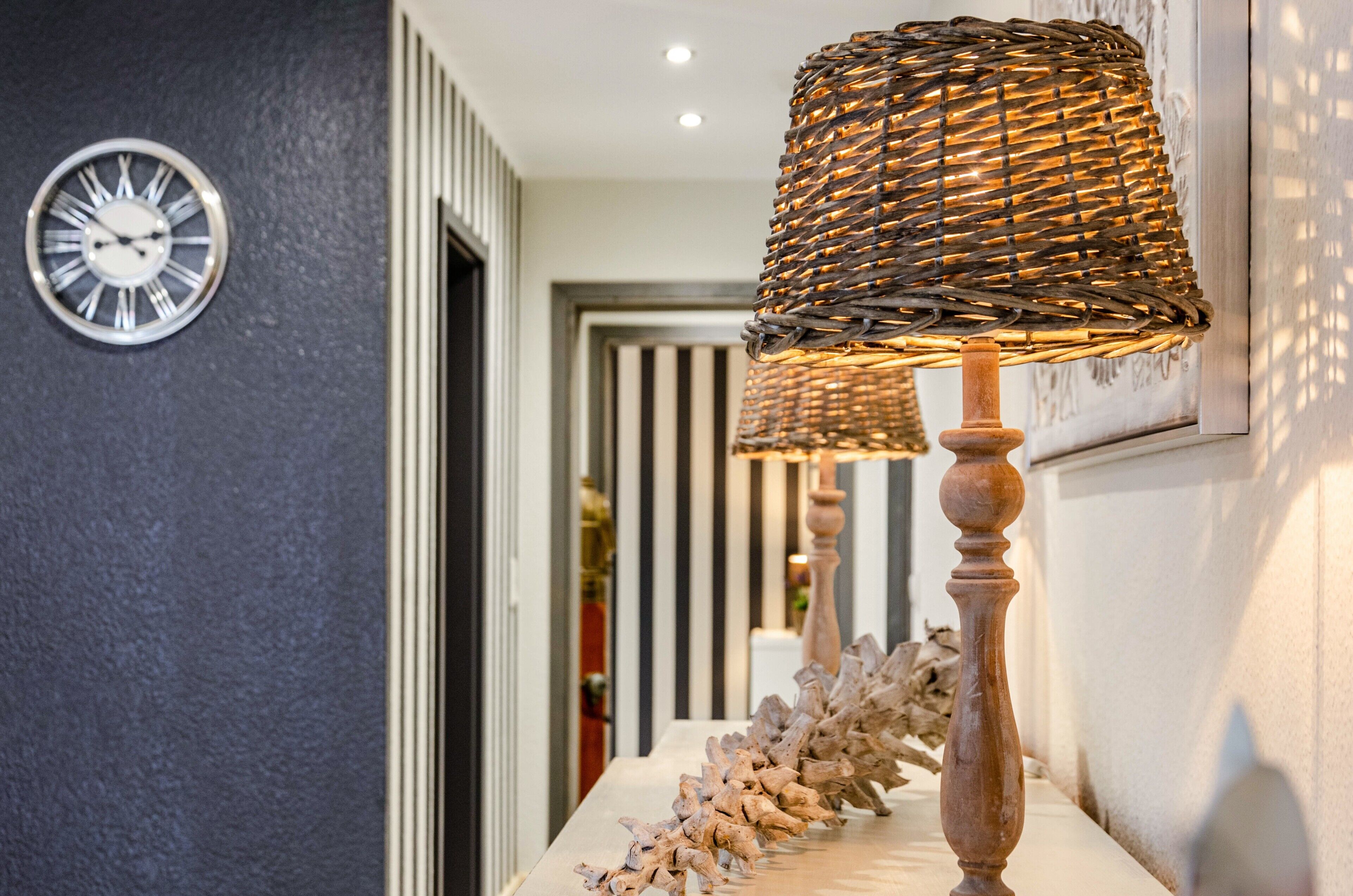 Een hal met een aan de muur bevestigd horloge, een rieten lamp en een decoratieve plantenstelling.