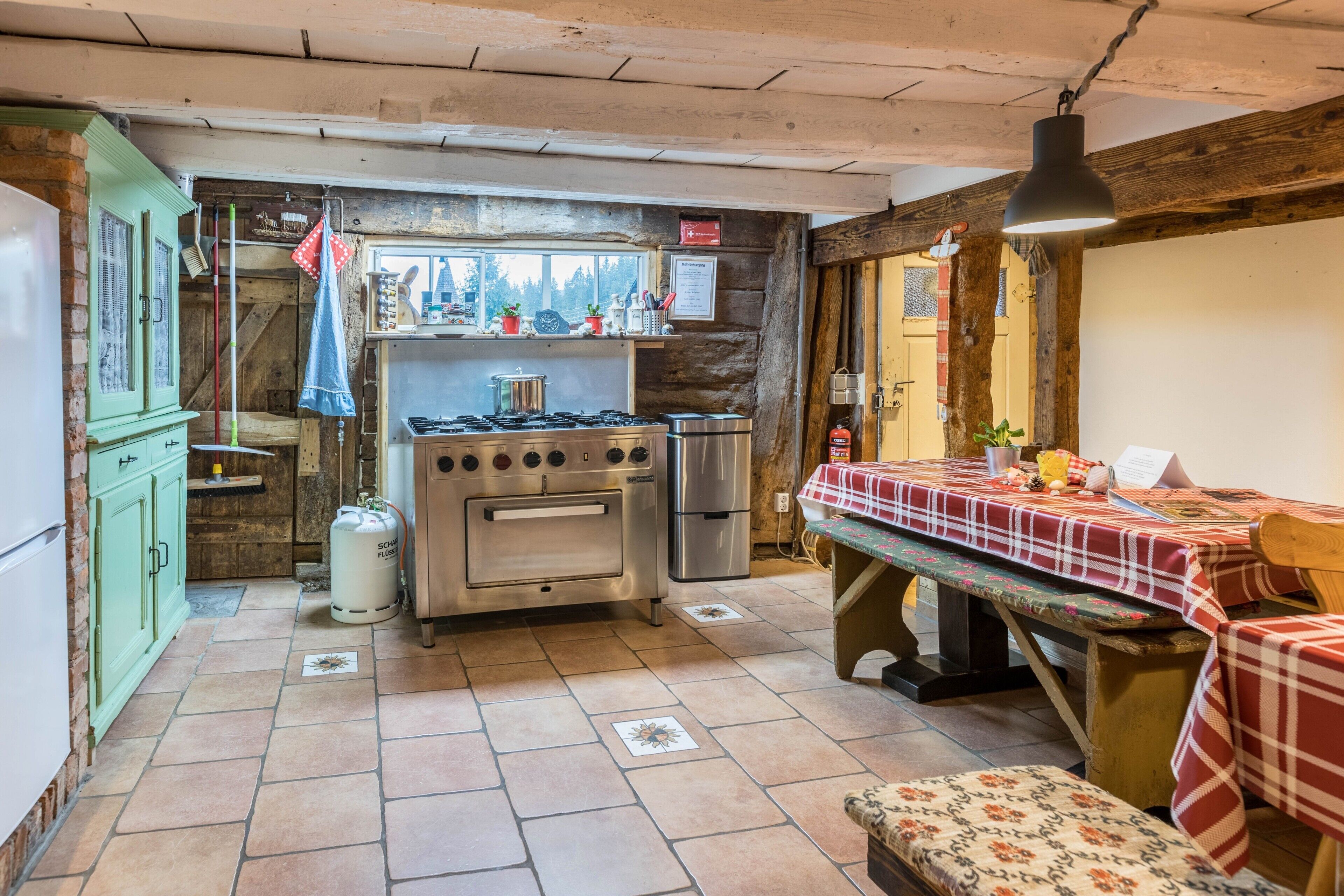 A rustic kitchen with a gas stove, oven, and refrigerator, a wooden bench, and a table with a checkered tablecloth.