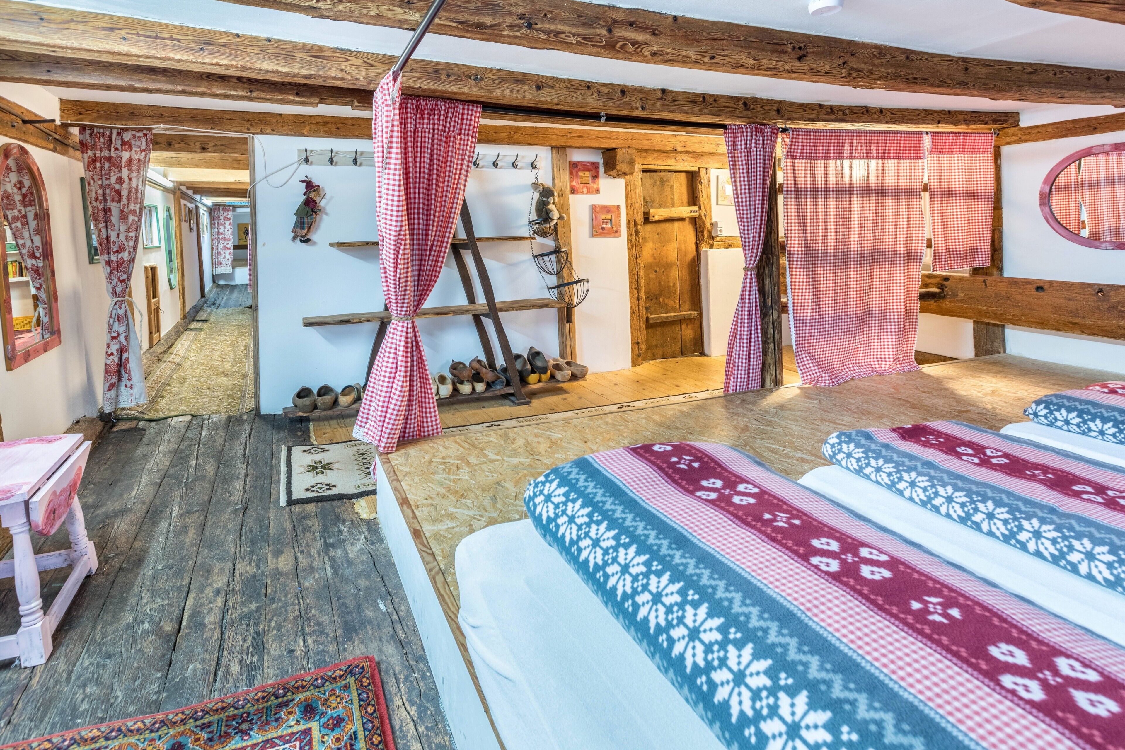 Ein rustikaler Raum mit Holzbalken, karierten Vorhängen, einem Bett mit gemusterten Textilien, einer Holzleiter und einem Wandregal mit Stiefeln.