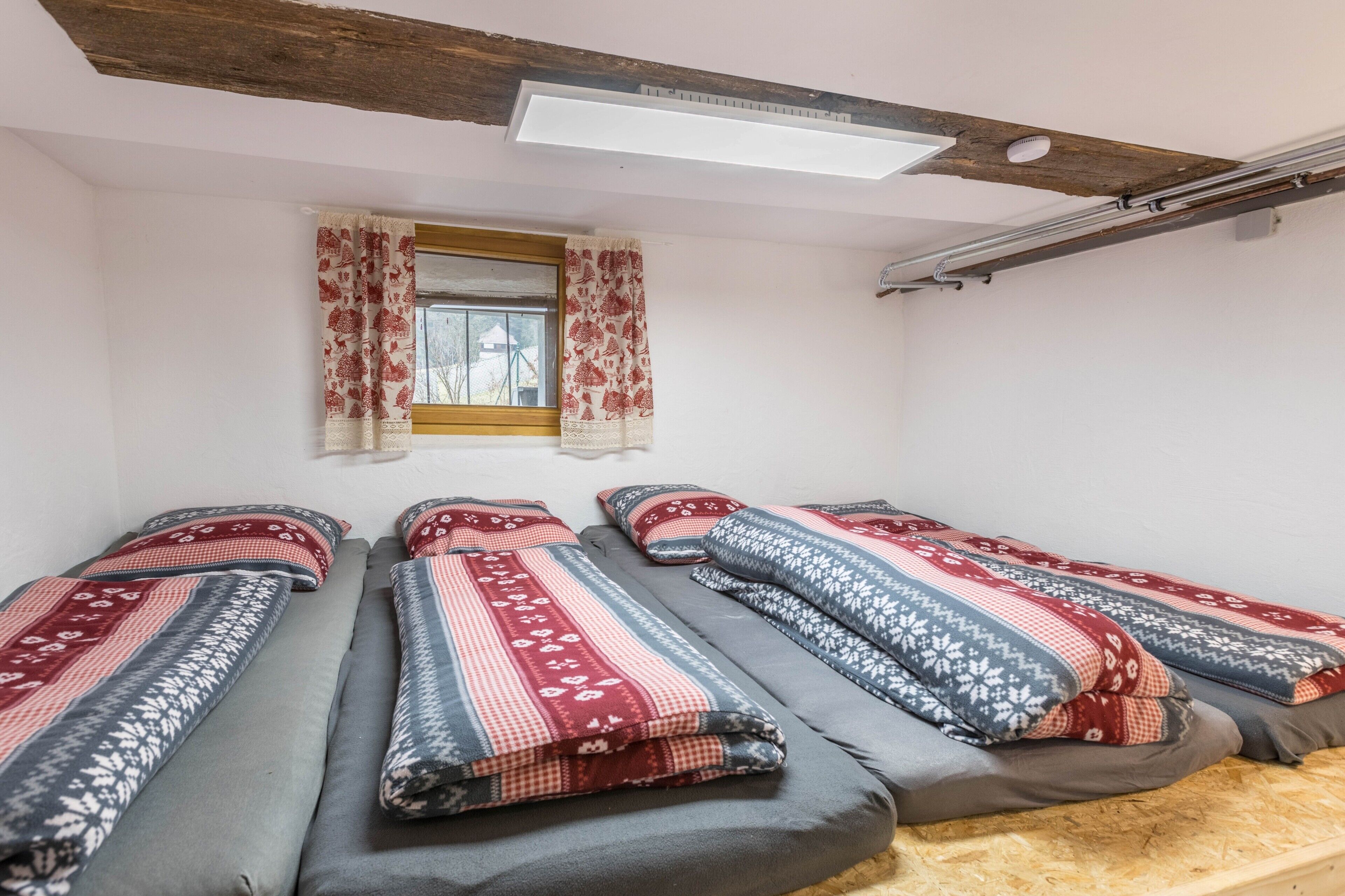 Une pièce avec trois lits, une fenêtre avec des rideaux, une poutre en bois au plafond et un luminaire suspendu au plafond.