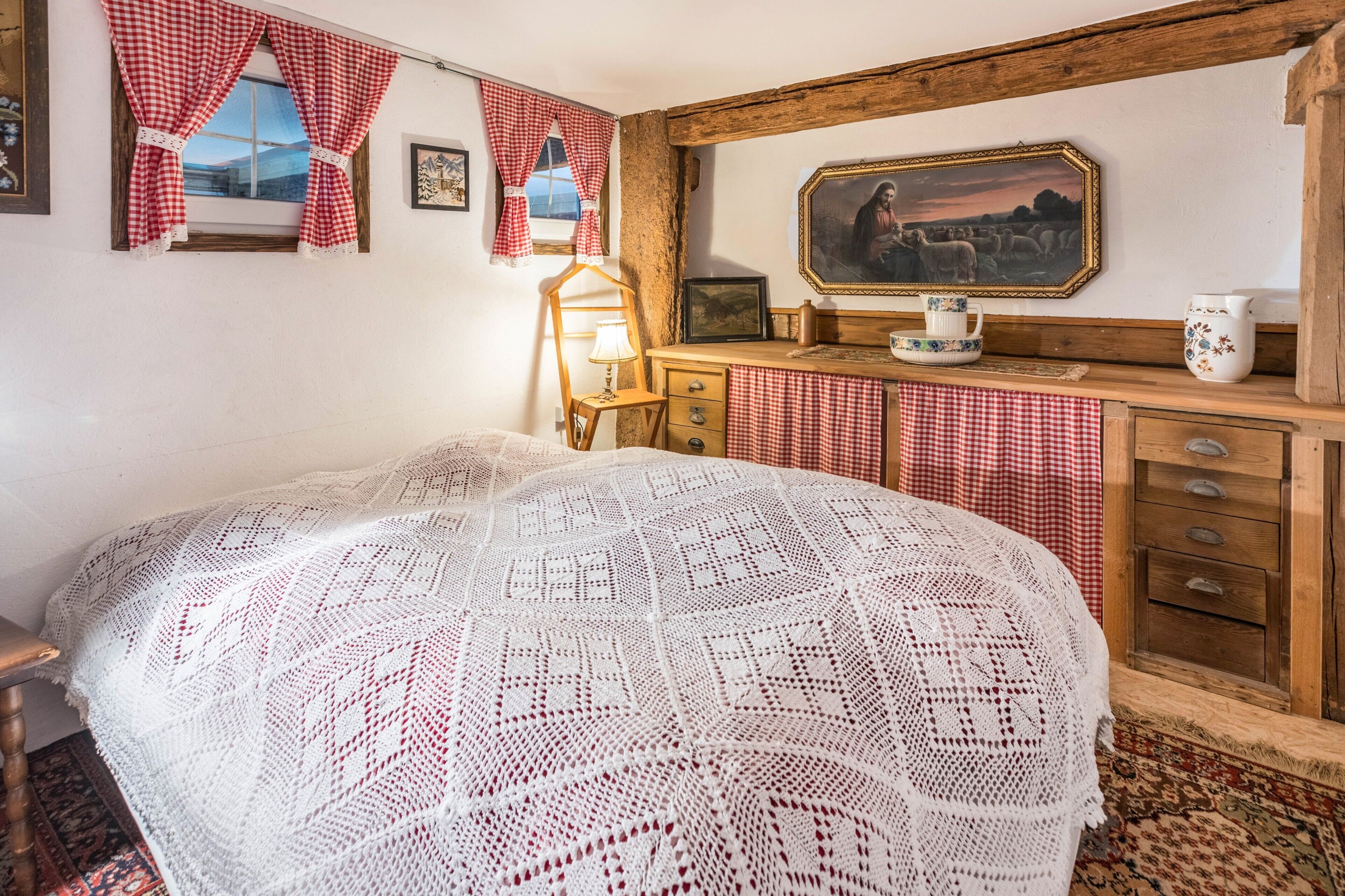 A room with a bed, a wooden dresser, a mirror, and a window with red and white checkered curtains.