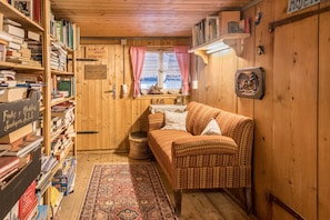 Ett mysigt rum med en soffa av rotting, en bokhylla i trä full med böcker, en mönstrad matta och ett litet fönster med röda gardiner.