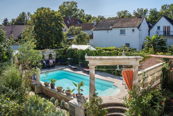 A swimming pool surrounded by greenery, a stone pillar with text, and a stone archway.