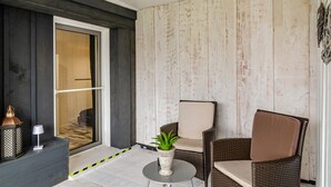 Een kamer met een donkere houten wand, een rieten stoel met kussen, een klein tafeltje met een plant en een glazen deur die naar een andere kamer leidt.