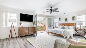 A bedroom with a bed, a ceiling fan, a TV, a dresser, a desk, and a balcony.