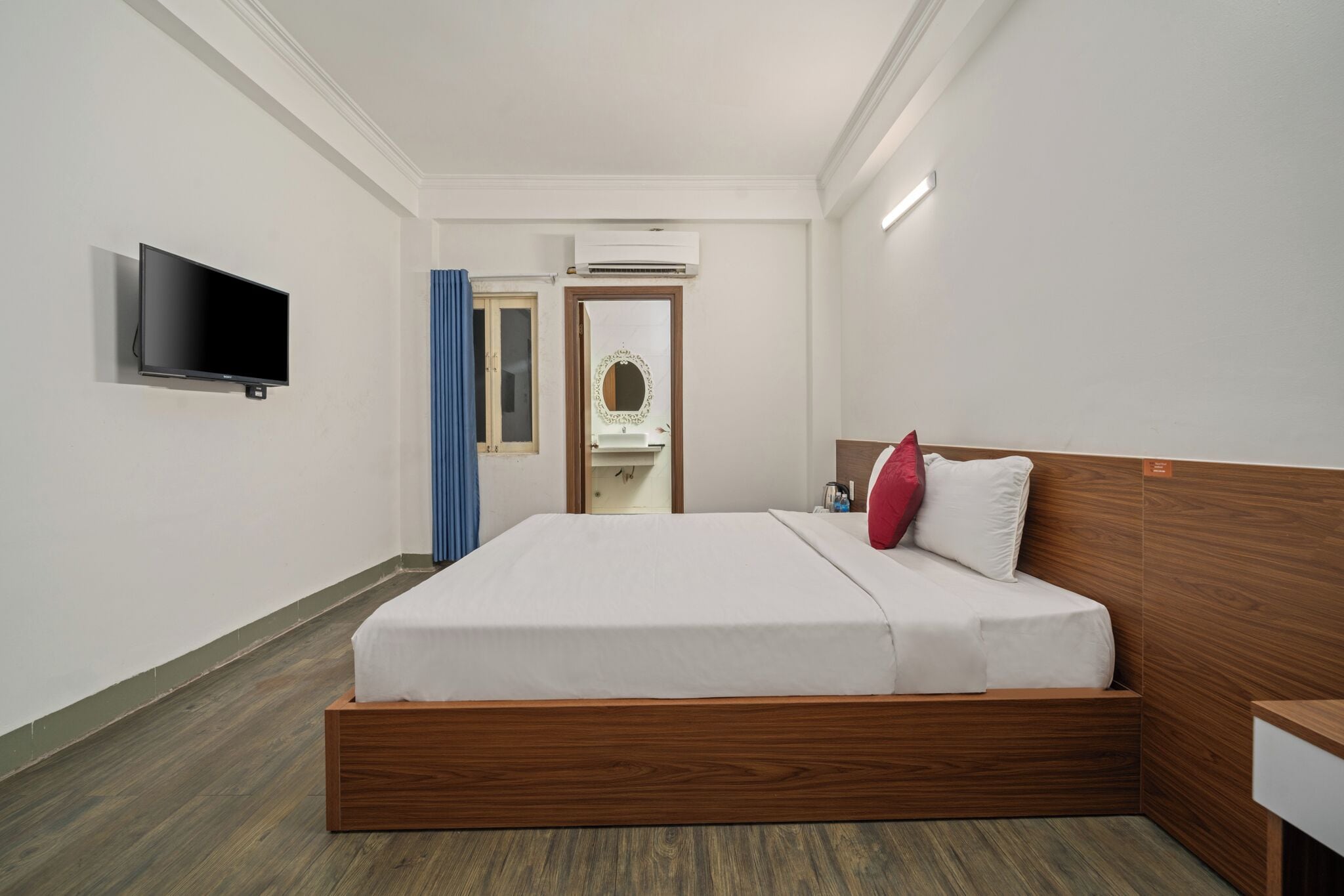 A modern hotel room with a wooden bed, a flat-screen TV, an air conditioning unit, and a mirror.