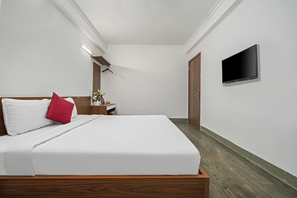 A neatly made bed with white bedding and a red pillow, a bedside table with a lamp, a small plant, and a television mounted on the wall.