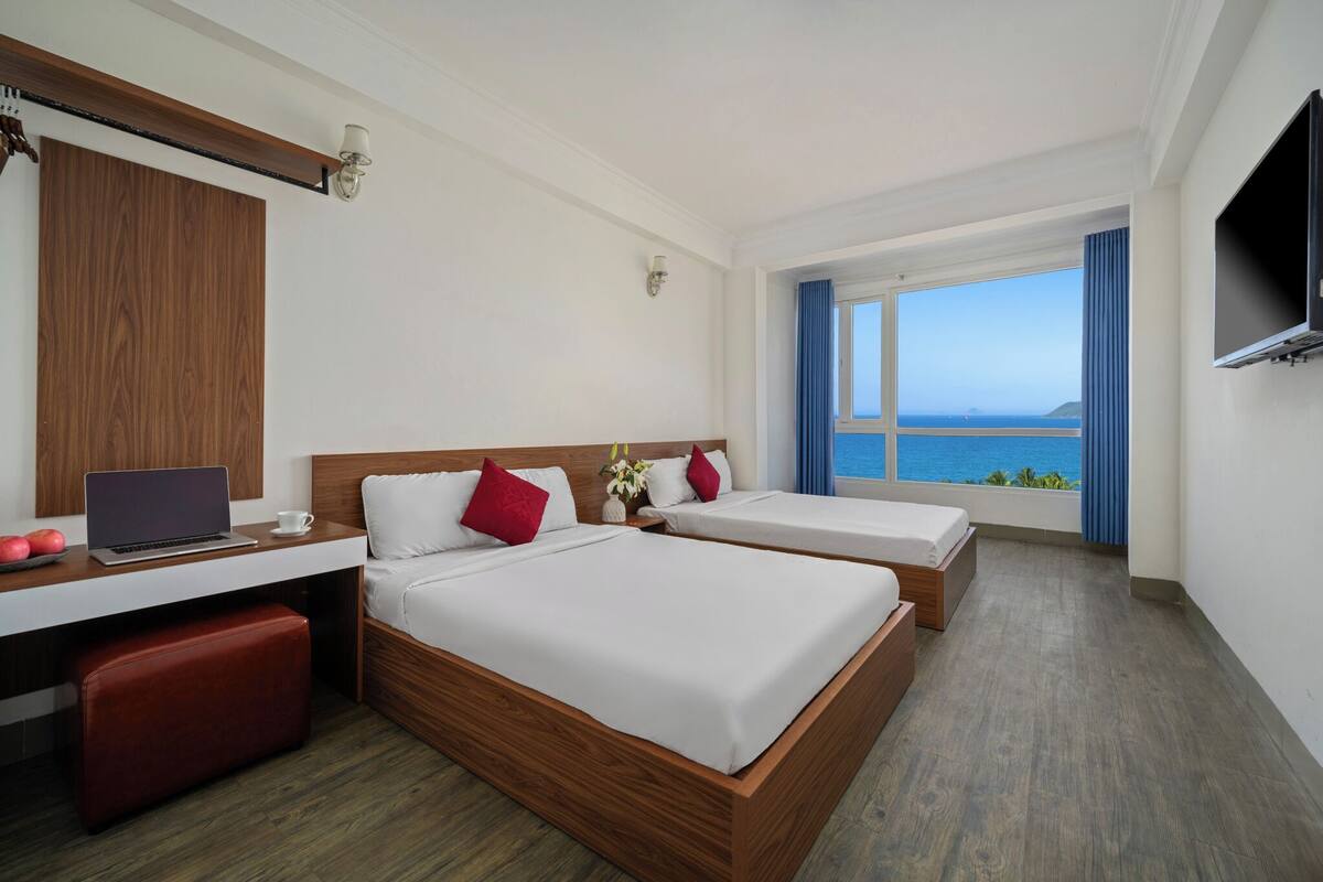 A hotel room with two beds, a desk with a laptop, a TV, and a large window with a view of the sea.