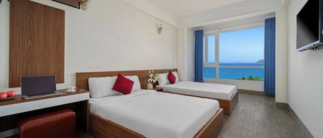 A hotel room with two beds, a desk with a laptop, a TV, and a large window with a view of the sea.