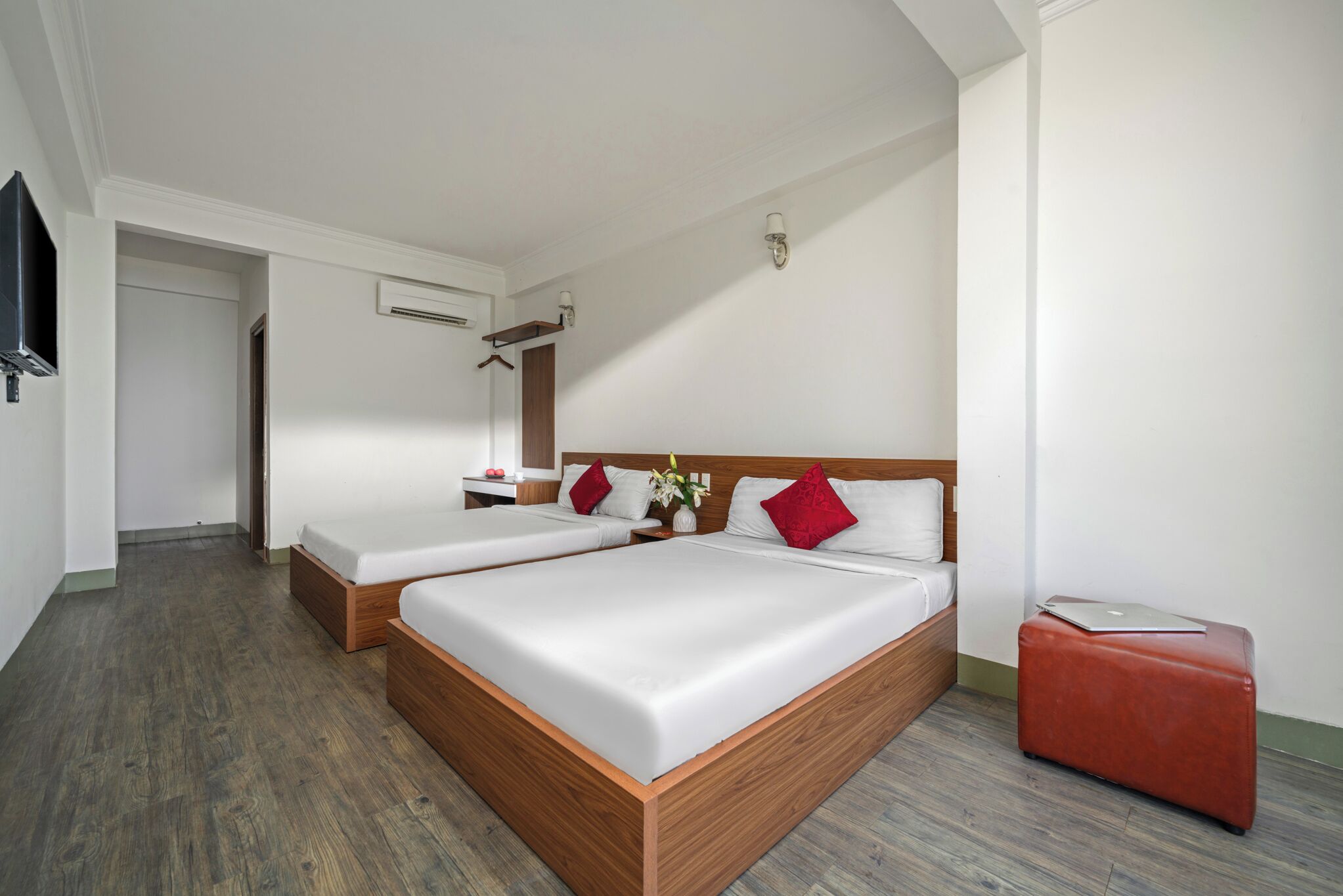Habitación de hotel con dos camas, cabecero de madera, maleta roja y televisor montado en la pared.