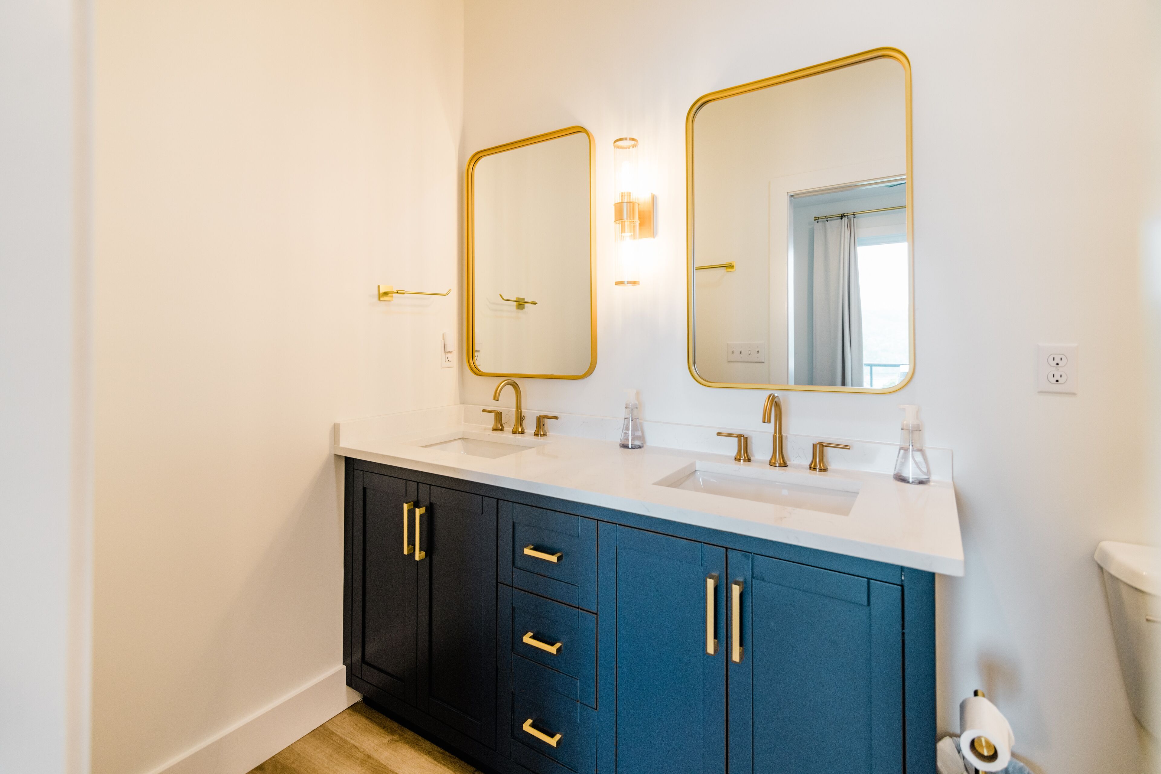 A modern bathroom with two sinks, dark blue cabinets, gold fixtures, and wall-mounted mirrors.