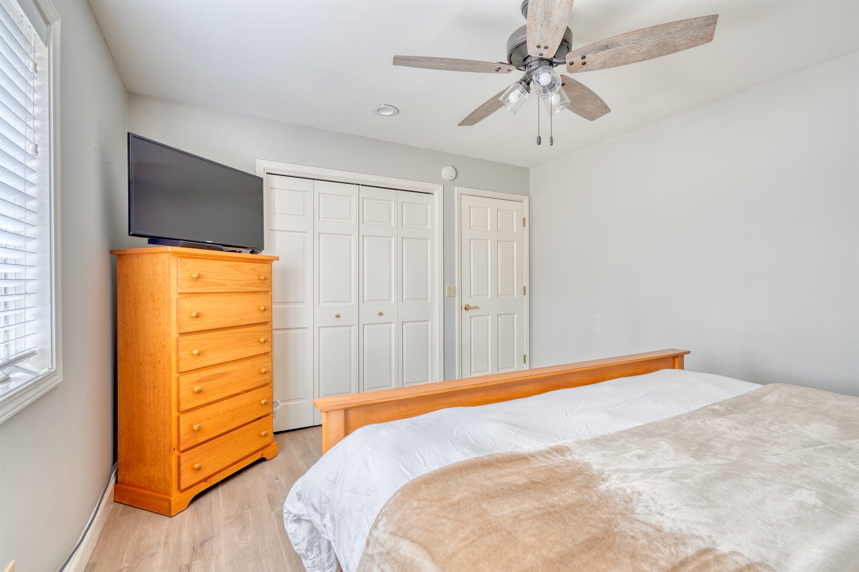 A bedroom with a bed, wooden dresser, television, white closet, and ceiling fan.