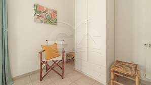 A room with a wooden chair, a small wooden stool, a wall with a floral painting, and a large white cabinet.