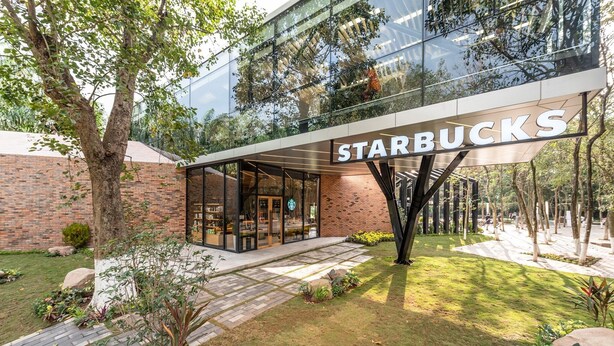 A Starbucks with a modern glass facade, outdoor seating, and a brick wall backdrop. - Friendly House QMP - Ecopark (Phung Cong)