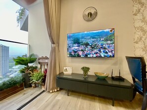 A modern living room with a large TV mounted on the wall, a console table with decorative items, and a view of a cityscape through a large window. - Friendly House QMP - Ecopark (Van Giang)
