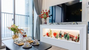A modern living room with a large flat-screen TV, a white fireplace with Christmas decorations, a dining table with ceramic items, and a window with curtains. - Friendly House QMP - Ecopark (Phung Cong)
