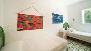 Quarto com cama, mesinha de cabeceira pequena, tapeçaria pendurada e janela com vista para o verde.