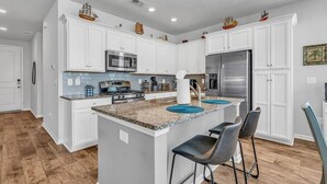 A modern kitchen with white cabinetry, a central island with bar stools, stainless steel appliances, and decorative items on the upper cabinets. - Stylish Beach Retreat | Close to Everything| 4 Bed (Myrtle Beach)