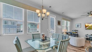 A dining area with a glass table surrounded by chairs, a chandelier, and a view of the outside through large windows with blinds. - Stylish Beach Retreat | Close to Everything| 4 Bed (Myrtle Beach)