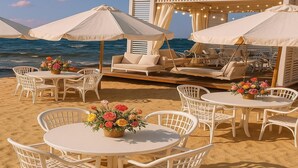 Un salon de plage doté de meubles en osier blancs, de tables rondes ornées de compositions florales et de grands parasols offrant de l’ombre.