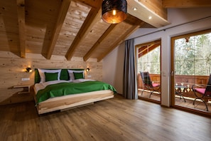 Un dormitorio de madera con una cama, una mesita de noche, una lámpara fijada a la pared y un amplio ventanal corredero que da a un espacio exterior con sillas.
