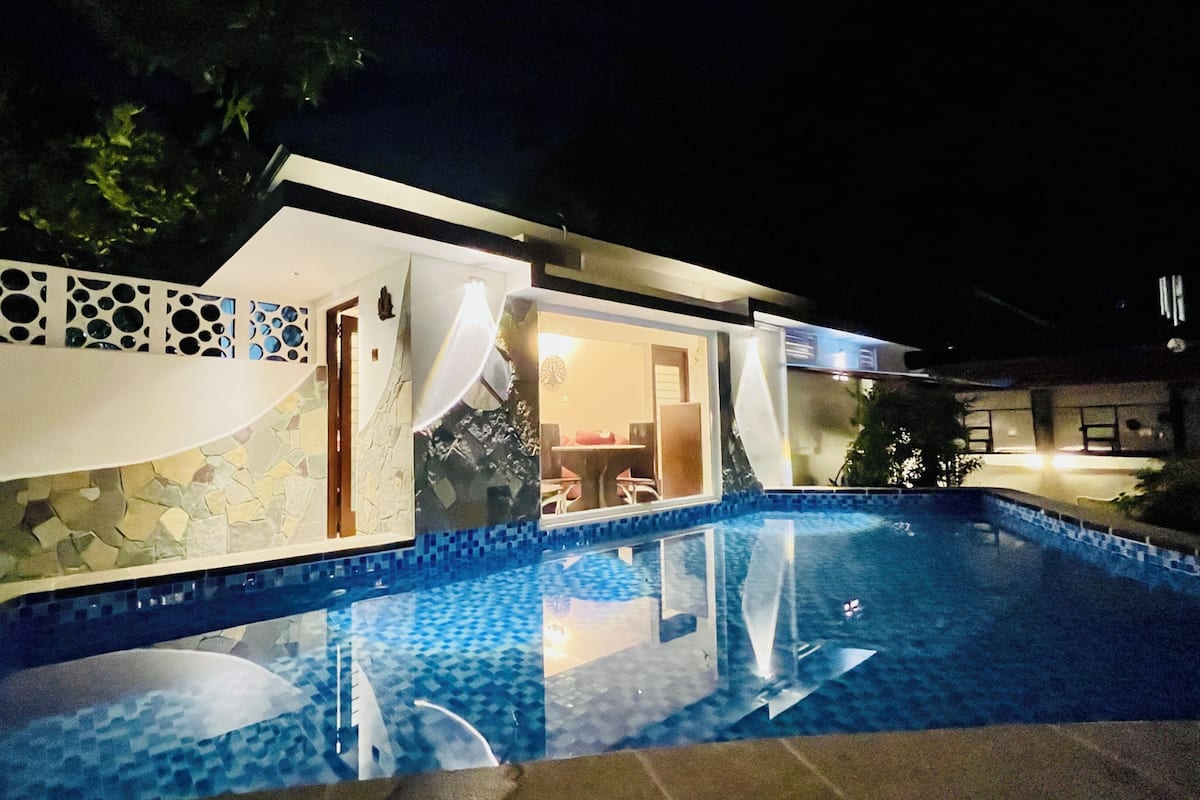 A modern house with a swimming pool, stone walls, and a dining area visible through the windows.